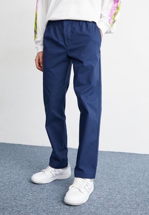Person trägt locker sitzende marineblaue Hose und weiße Sneakers, steht auf einem grau geteppten Boden vor einem schlichten weißen Hintergrund.
