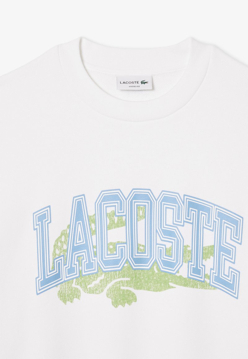 T-shirt bianca con il testo "LACOSTE" azzurro chiaro e un grafico di coccodrillo verde. Collo a giro, vestibilità ampia e texture del tessuto morbida.