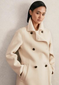 Beige dvojsrdcový kabát s vysokým golierom, vyrobený z jemného materiálu. Má tmavé gombíky a bočné vrecká, ktoré zdôrazňujú štruktúrovaný dizajn.