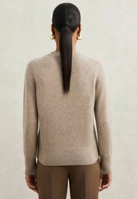 Pulover bej tricotat, cu mâneci lungi, mansete și tiv cu dungi. Textură moale, fără modele. Părul este prins înapoi; poartă cercei rotunzi din aur.