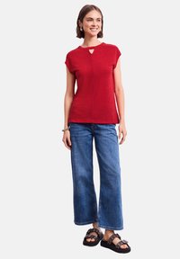 Maglietta rossa lavorata a maglia con maniche corte e dettaglio a chiave sul collo, abbinata a jeans blu a gamba larga e sandali neri con hardware decorativo.