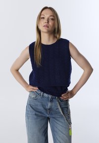 Young woman with long blonde hair wearing a sleeveless navy knit top and loose light blue jeans with chains on the right pocket.