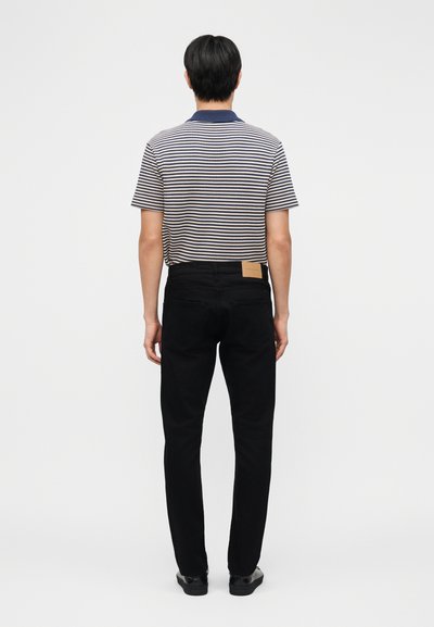 Mees seisab seljaga vaataja poole, kandes lühikeste varrukatega mereväe- ja valge triibulist särki, musti pükse ja musti kingi valgel taustal.