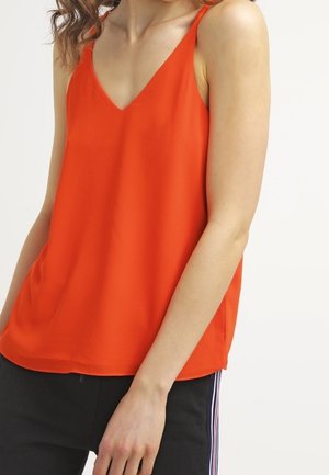 Blusa rossa senza maniche con scollo a V, texture liscia e vestibilità ampia. Abbinata a pantaloni scuri con strisce laterali.