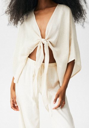 Femme portant un haut ample crème noué devant avec de larges manches et un pantalon assorti taille haute à cordon, debout devant un fond uni.