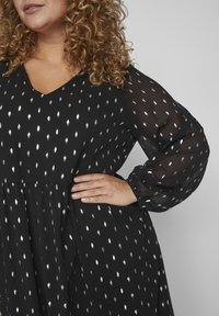 Vestido negro con escote en V, con estampados de puntos plateados, mangas largas transparentes y una silueta fluida con textura fruncida en la cintura.