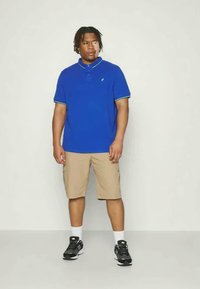 Camisa polo azul com colarinho contrastante, calções cargo bege e sapatos desportivos pretos com solas brancas, em pé sobre um fundo neutro.