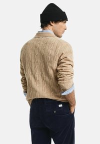 Man in een beige kabelgebreide trui over een blauwe shirt, marineblauwe corduroy broek en een zwarte gebreide beanie, zijwaarts gekeerd met de handen in de zakken.