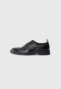 Svart lær vinge-tips oxford-sko med brogue-detaljer, grepstropp på hælen og tykk gummisåle mot en hvit bakgrunn.