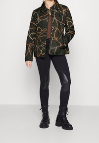 Grön vadderad jacka med mönster av guldkedjor, tryckknappar och krage. Matchad med svarta leggings med läderdetaljer och svarta snörboots.