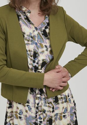 Person wearing a green open-front cardigan over a multi-colored abstract patterned buttoned dress, hands clasped at the waist.