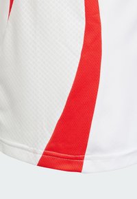 Sportovní dres s bílým tělem a červeným diagonálním pruhem. Vyrobený z prodyšného materiálu s texturovaným povrchem a zesíleným šitím.