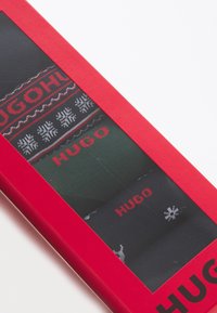 Tre par sokker i en rød rektangulær kasse. To par er sorte med røde detaljer; det ene har et snefnugmønster. Grønne par med rødt logo.