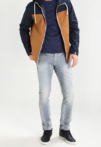 Homme portant une veste à capuche bleu marine et beige par-dessus une chemise bleu marine, un jean bleu clair et des baskets noires avec des semelles blanches, debout devant un fond blanc.