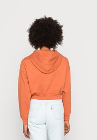 Cropped hoodie in coral orange, made of soft fabric with a rounded hood and ribbed cuffs. Paired with light blue high-waisted jeans.
