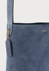 Niebieska torba na ramię z zamszu, z zawieszką ze skóry i złotym łańcuszkiem, z małym złotym logo "Coach New York" blisko górnej krawędzi.