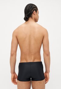 Un maillot de bain noir avec un design lisse et près du corps. Le dos du modèle est montré, mettant en valeur le style minimaliste du vêtement et ses lignes épurées.