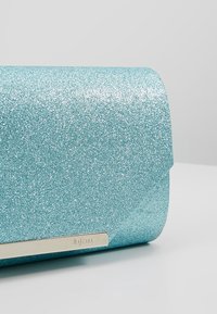 Pochette à paillettes bleues étincelantes avec rabat arrondi et accent en métal argenté gravé de "Mascara" sur une surface grise claire uniforme.