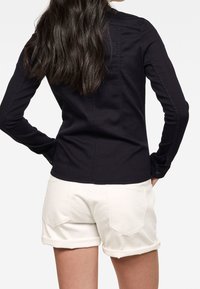 Camicia a maniche lunghe color blu navy con design aderente, polsini con bottoni e dettagli cuciti, abbinata a pantaloncini bianchi risvoltati con tasche posteriori.