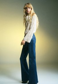 Jeans in denim blu a zampa d'elefante con vita alta, abbinati a un top lungo a maniche lunghe di tessuto leggero e testurizzato. Il tessuto morbido e la vestibilità comoda creano un look rilassato.