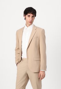 Costume beige à la coupe ajustée ; revers crantés, fermeture à un bouton et une texture subtile. Porté sur une chemise blanche à manches longues.