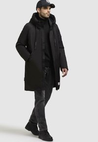 Sort isoleret parka med hætte, lynlås foran og snore. Kombineret med sorte jeans og sorte sneakers, hvilket skaber et monokromt look.