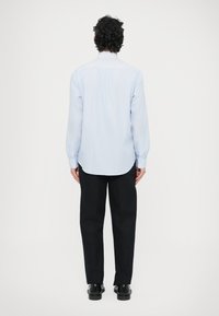 Jasnoniebieska, paskowana koszula z długimi rękawami i zaokrąglonym dołem, zestawiona z czarnymi spodniami i czarnym obuwiem, widziana od tyłu.