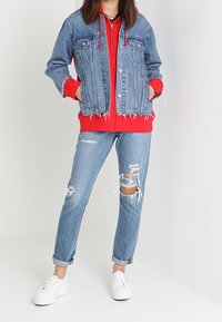 Džínová bunda s opranými okraji, navršená přes červenou mikinu s kapucí. Nošeno s roztrhanými světle modrými džínami a bílými teniskami.