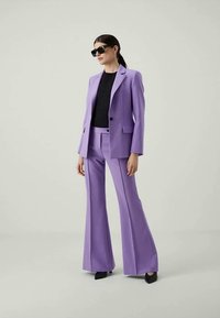 Woman wearing black sunglasses, black top, and a lilac suit with flared trousers and black pointed shoes, standing against a plain background.