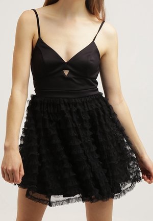 Femme portant une robe noire sans manches avec un corsage ajusté et une jupe à volants superposés, debout devant un fond uni.