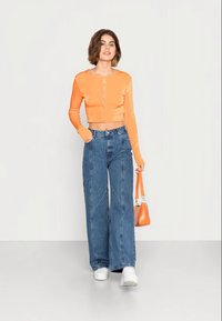 Top laranja cropped de mangas longas com botão à frente, combinado com jeans de ganga azul de perna larga e alta, e ténis brancos, segurando um saco laranja.