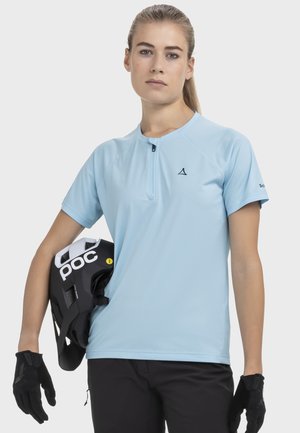 Junge Frau in hellblauem Sportshirt mit kurzen Ärmeln, schwarzen Handschuhen und schwarzer Hose, hält einen schwarz-weißen Fahrradhelm seitlich.