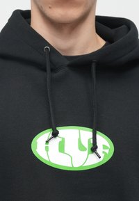 Prim-plan al unei persoane care poartă un hanorac negru cu un logo oval alb și verde și sfori atârnând în față.