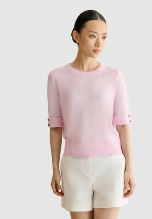 Mujer que lleva un suéter rosa claro de manga corta con detalles de botones y shorts blancos de talle alto sobre un fondo sencillo.
