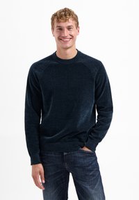 Marineblauwe trui van een zachte, gestructureerde brei met een ronde hals en geribde manchetten, gecombineerd met donkere spijkerbroeken. Model met een glimlach.