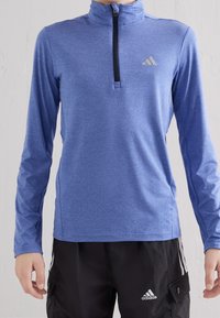 Persona che indossa un top sportivo a maniche lunghe blu con zip e pantaloni neri Adidas con strisce e loghi bianchi.