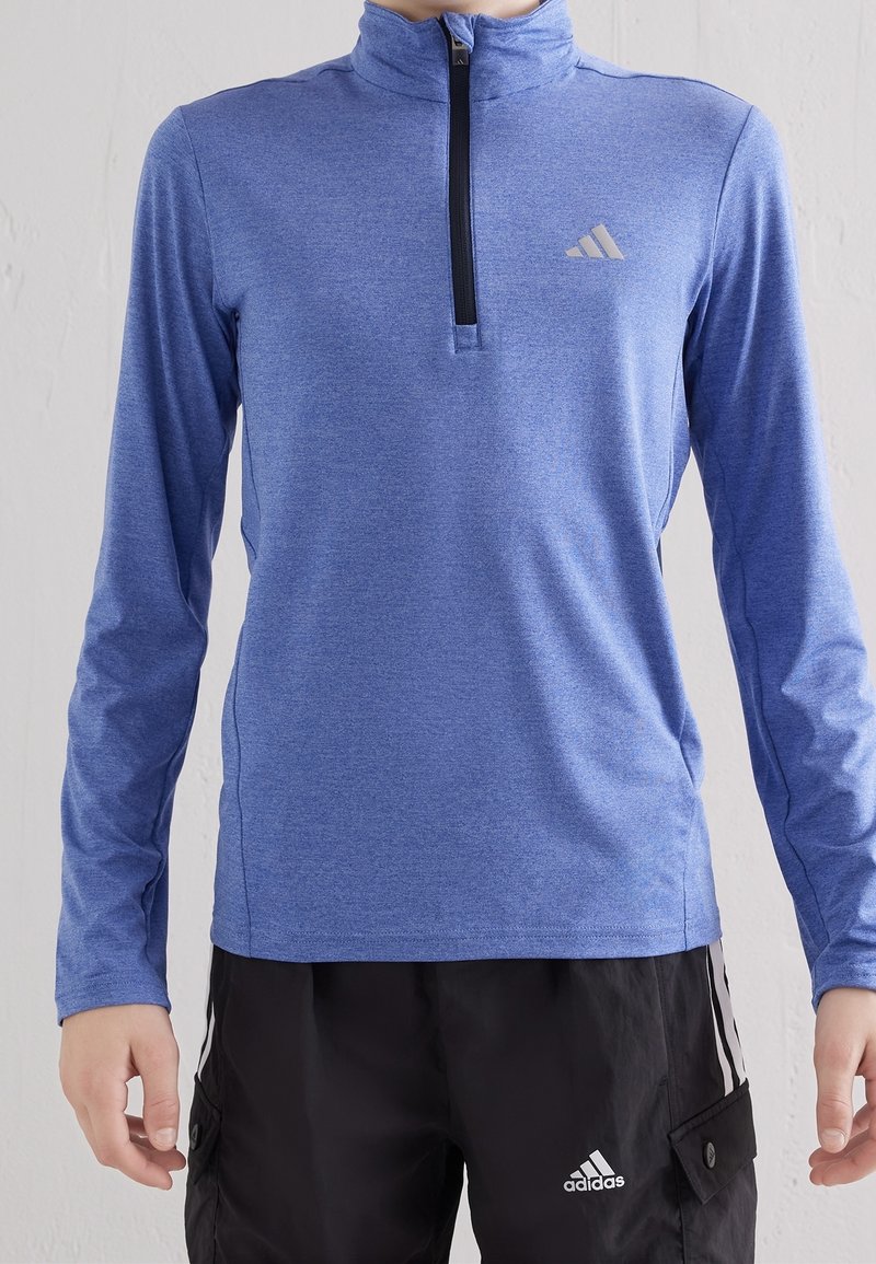 Persona che indossa un top sportivo a maniche lunghe blu con zip e pantaloni neri Adidas con strisce e loghi bianchi.