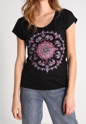 Vrouw draagt een zwart shirt met korte mouwen met een roze en paarse cirkelvormige mandala met olifanten, gecombineerd met een blauwe spijkerbroek.