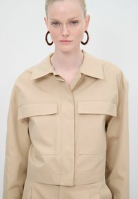 Giacca a camicia cropped beige con colletto strutturato, due tasche frontali e chiusura a bottoni. Tessuto liscio e design minimalista.