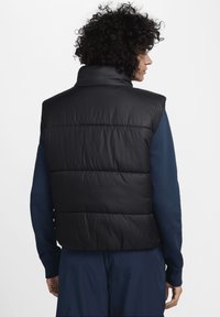 Μαύρο αμάνικο μπουφάν με ψηλό γιακά, καπιτονέ σχέδιο και ελαστικό τελείωμα. Φοριέται πάνω από ένα σκούρο μπλε μακρυμάνικο πουκάμισο, με ομαλή υφή.