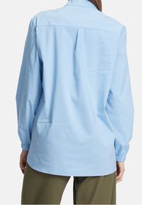 Jasnoniebieska koszula z długim rękawem ze ściąganymi mankietami noszona przez osobę stojącą, pokazana z tyłu, połączona z oliwkowymi spodniami.