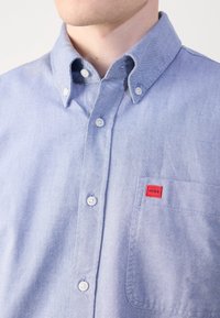 Gros plan d'une chemise bleu clair à boutons blancs avec une petite étiquette rouge "HUGO" sur la poche poitrine.