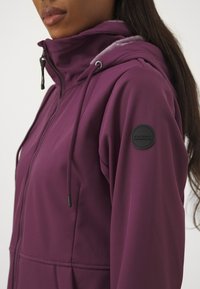 Fleece-gevoerde, rits-jack in diep paars, met een hoge kraag, een trekkoordcapuchon en een ronde zwarte logo-patch op de mouw.