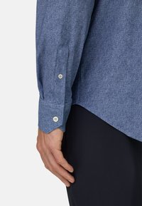 Blauw gemêleerde overhemdstof met een knoopmanchet. Heeft een licht gebogen zoom en een gestructureerd oppervlak. Close-up van een hand op donkere kleding.