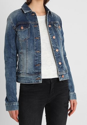 Femme portant une veste en jean bleue avec des boutons en cuivre sur un haut blanc et un jean noir, debout devant un fond gris clair uni.