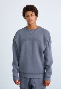 Grå hettegenser med rund hals og brodert "ALPHA INDUSTRIES"-tekst. Har en avslappet passform, ribbede mansjetter og slitt detaljer.