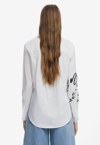 Camicia bianca a maniche lunghe con orlo curvato e un motivo astratto nero sulla manica sinistra. Il tessuto appare liscio e leggero.