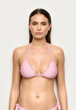 Mujer con cabello castaño liso que lleva un top de bikini rosa y blanco a rayas con bordes fruncidos, de pie contra un fondo claro y sencillo.