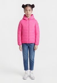 Fata cu două cocuri, purtând o jachetă matlasată roz aprins, blugi albaștri și adidași albi înalți, stând în picioare pe un fundal simplu.