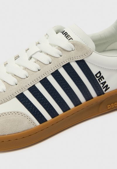 Hvide og beige sneakers med marineblå striber, tekstureret ruskind og gummisåler. Har snørebåndsdesign og "DEAN" trykt på siden.
